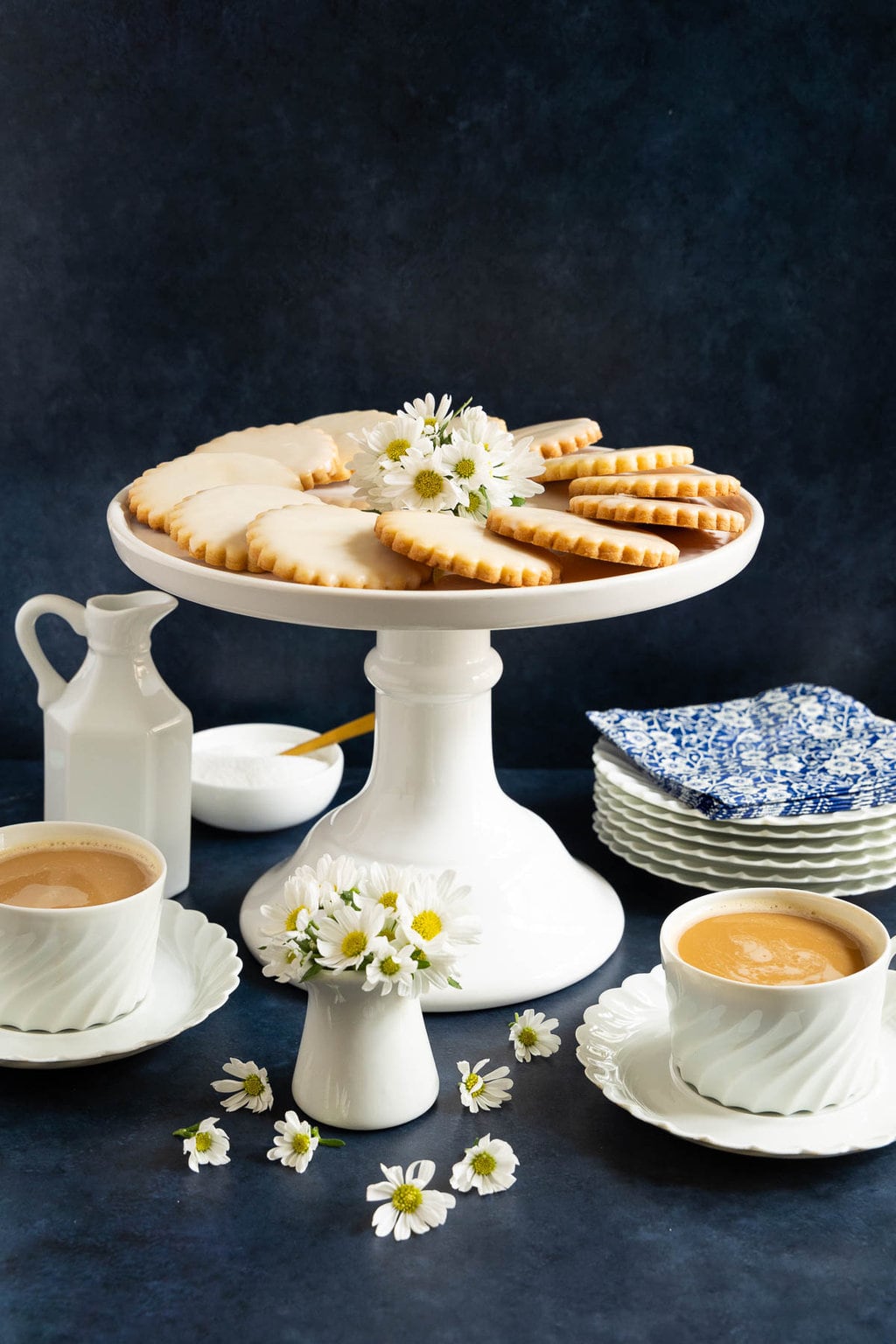 Vertical photo of a batch of Lemon-Glazed French Sable Cookies on a white pedestal serving plate with cups of coffee.