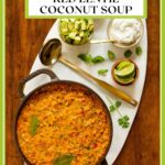 Vertical overhead photo of a cast iron pot of Curried Red Lentil Coconut Soup with small bowls of avocado wedges, lime, and fresh basil and cilantro leaves.
