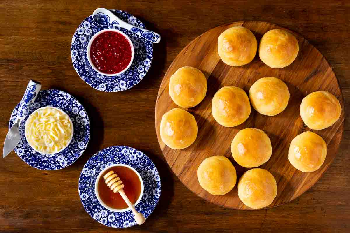 Horizontal overhead photo of Easy No-Knead Potato Rolls on a round wood platter with cups of jam, honey and butter.