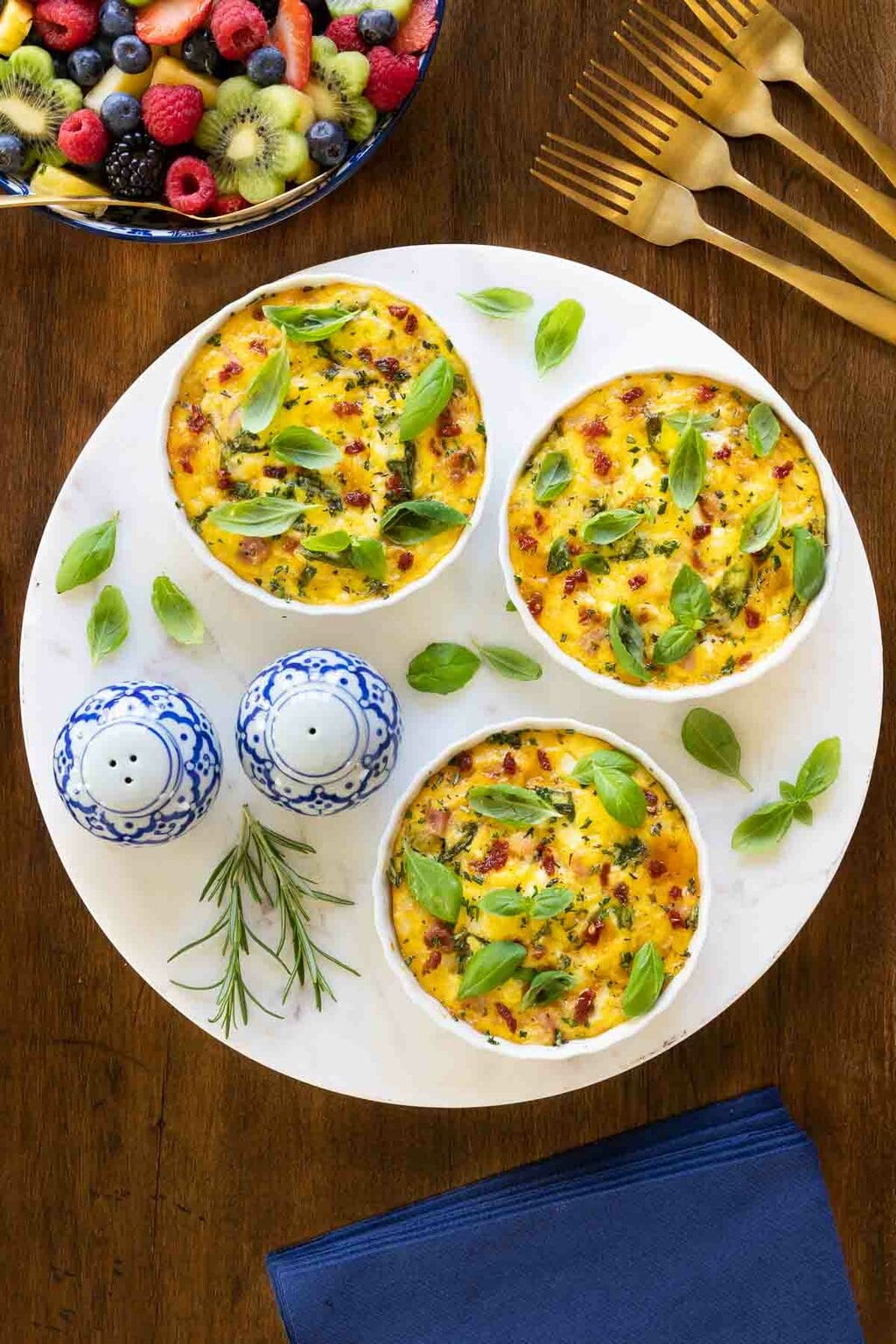 Vertical overhead photo of three Fresh Herb and Ham Crustless Quiches in individual serving ramekins and garnished with fresh basil and rosemary.