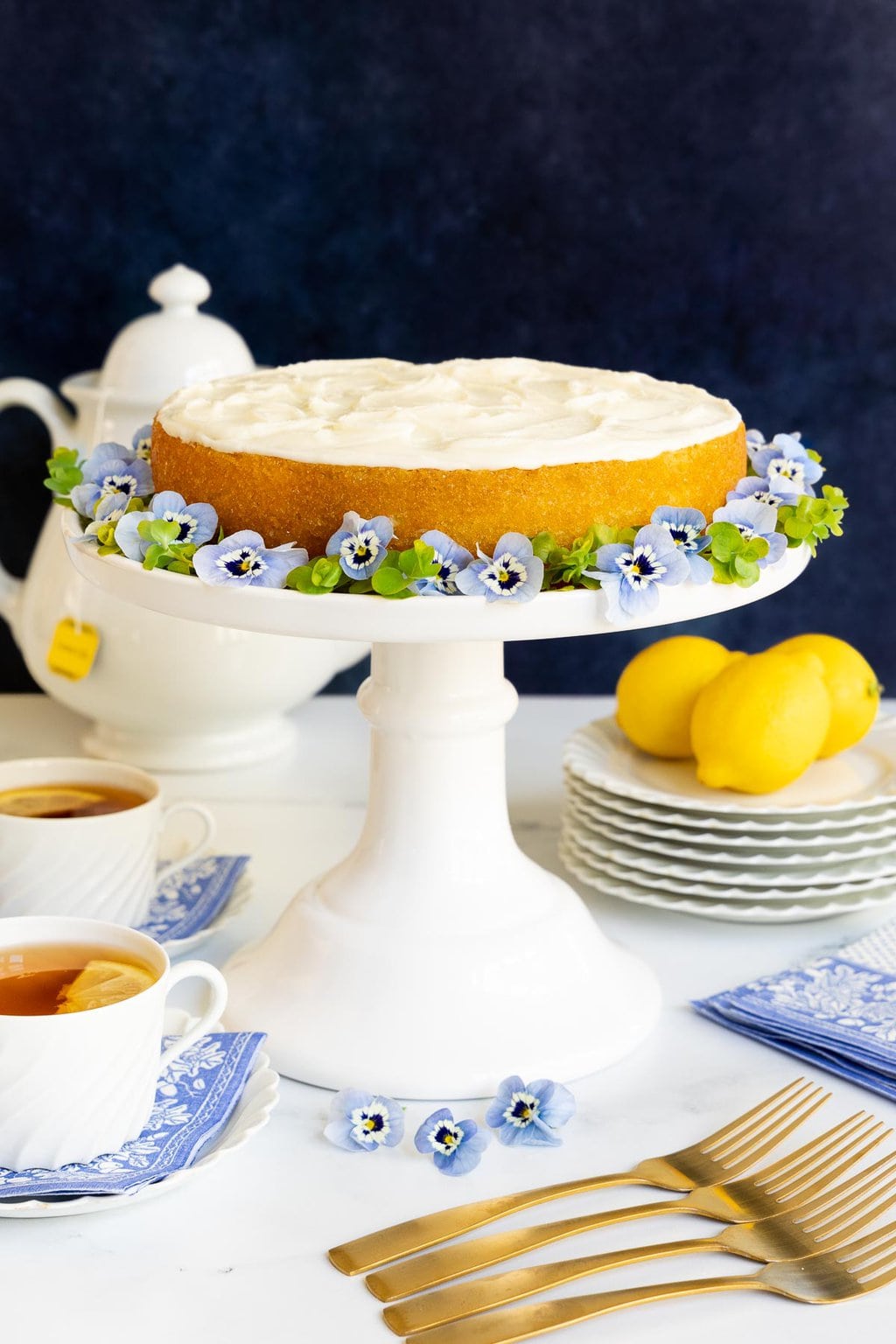 Vertical photo of a Ridiculously Easy Lemon Buttermilk Cake on a tall white pedestal serving plate. The cake is surrounded by delft blue pansies.