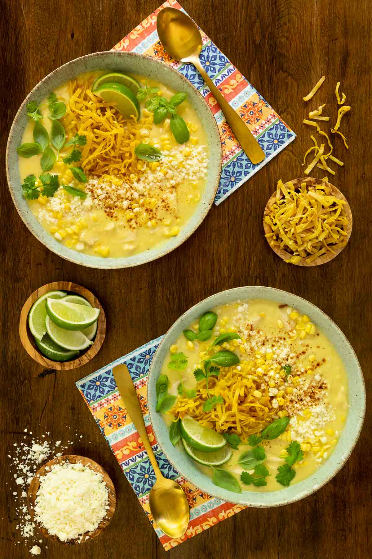 Vertical overhead photo of two bowls of Mexican Street Corn Chicken Soup garnished with tortilla strips, basil leaves cheese and lime wedges.