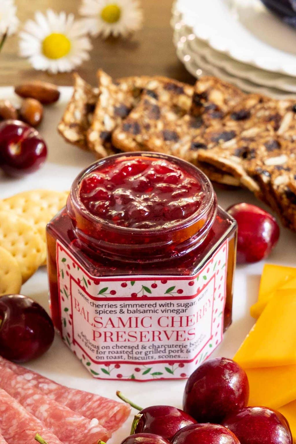 Vertical photo of a jar of Balsamic Sweet Cherry Preserves on a charcuterie board with a custom label for gift giving.