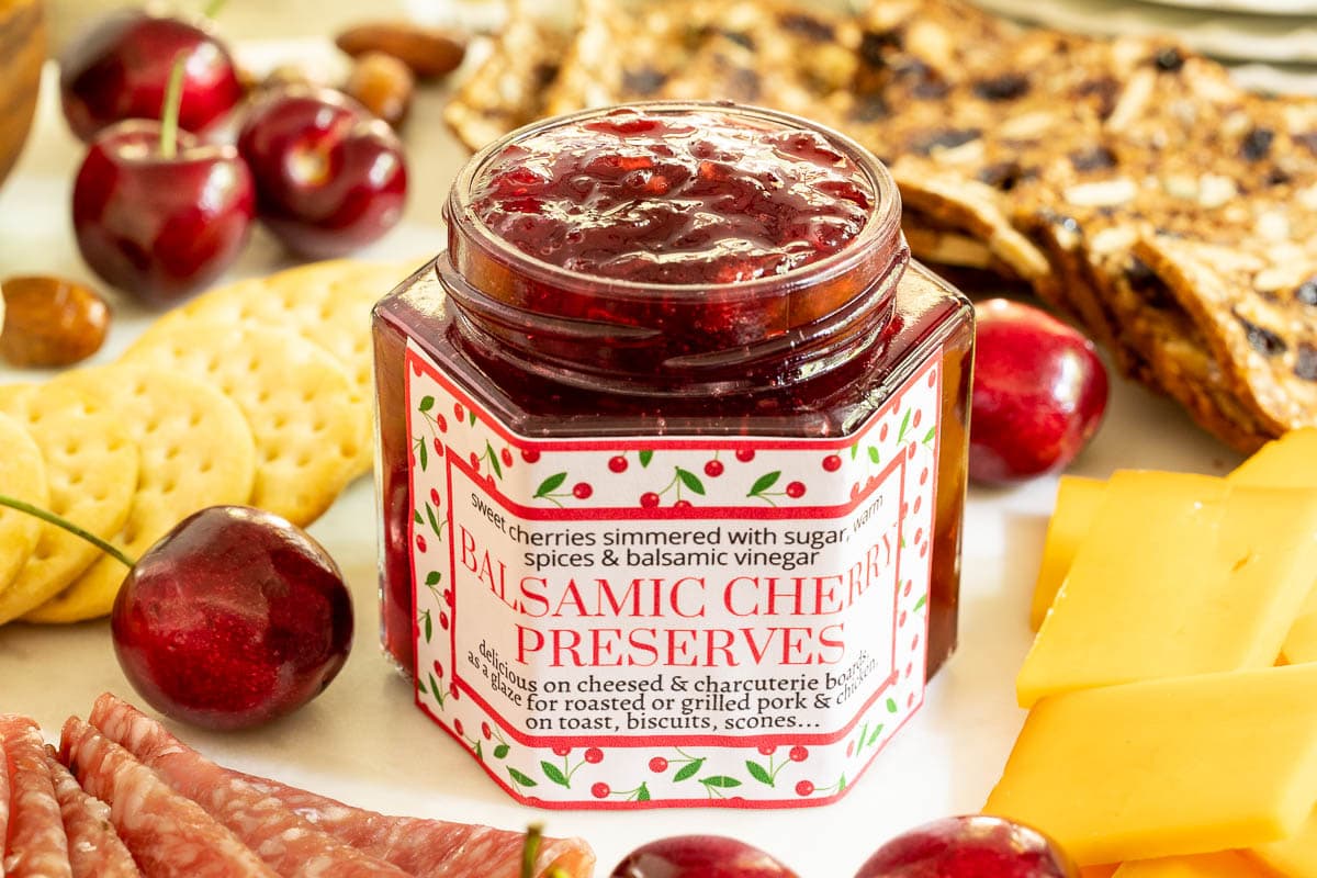 Horizontal closeup photo of a jar of Balsamic Sweet Cherry Preserves on a charcuterie board with a custom label for gift giving.