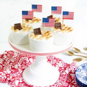 Horizontal photo of a batch of Easy Brownie S'Mores in Ramekins on a white pedestal serving plate decorated for the Fourth of July with American flags.