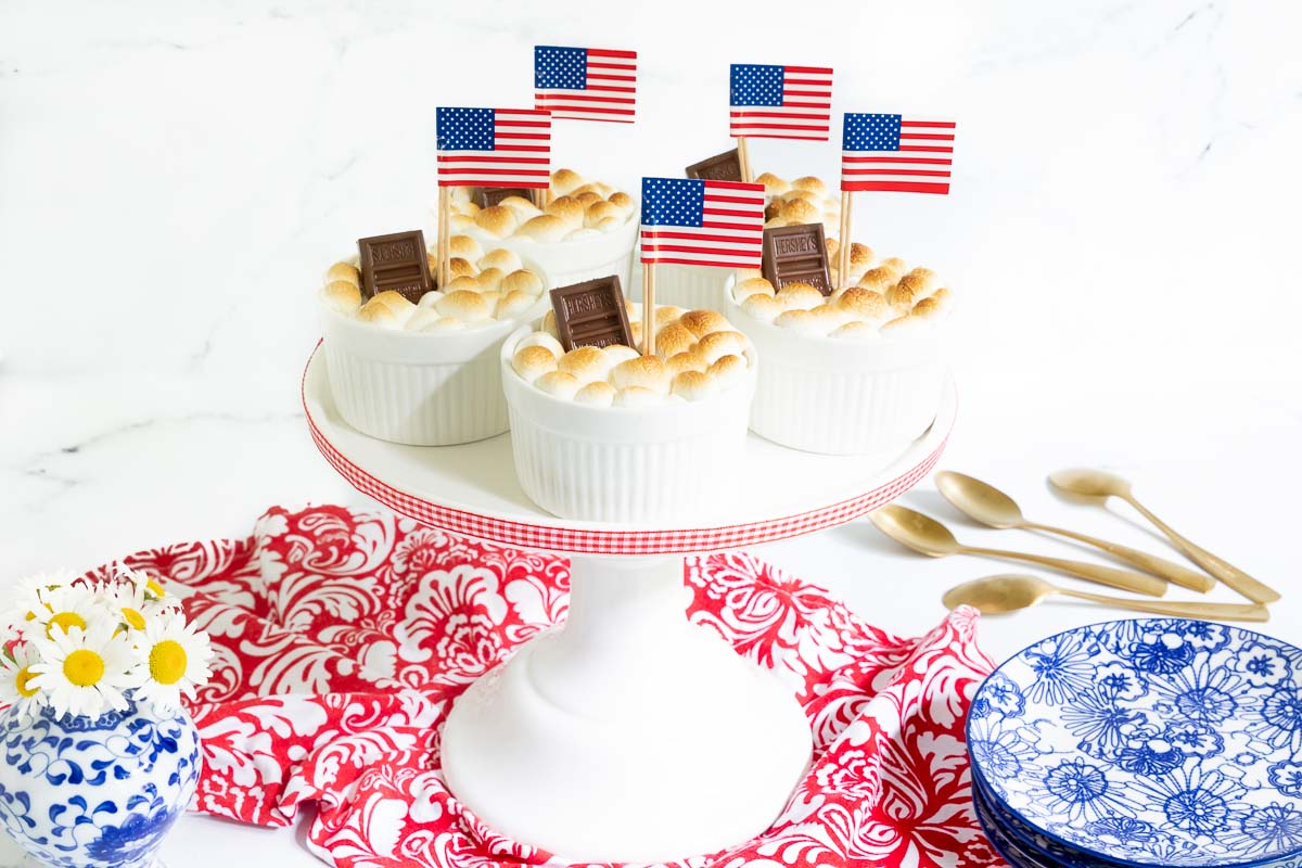 Horizontal photo of a batch of Easy Brownie S'Mores in Ramekins on a white pedestal serving plate decorated for the Fourth of July with American flags.