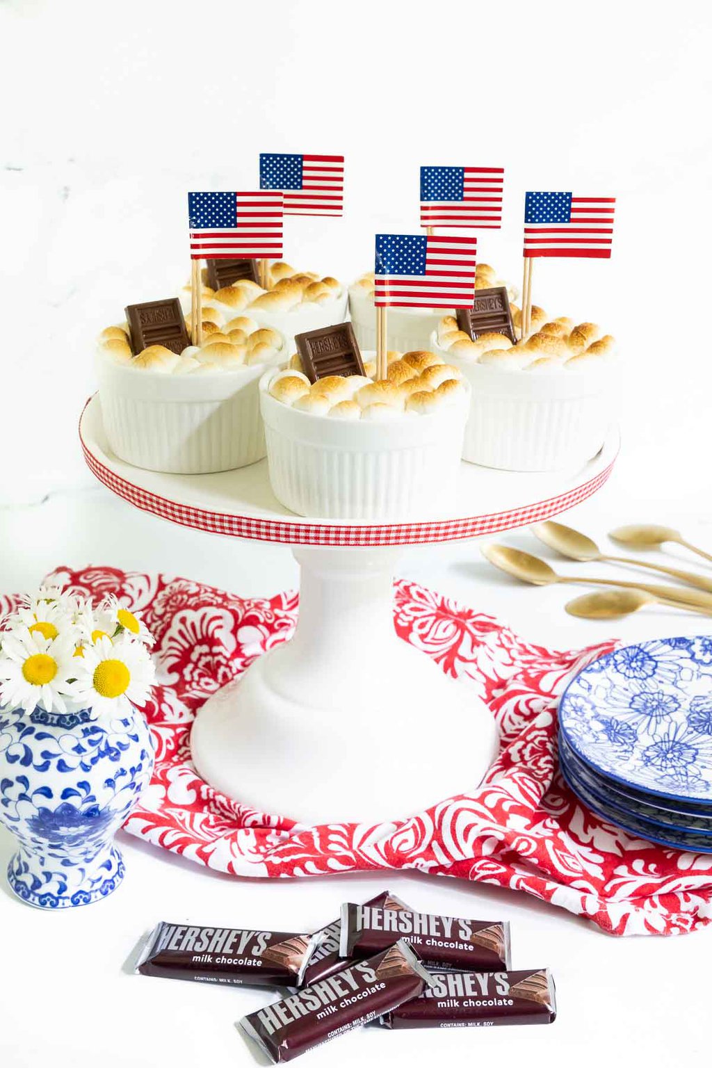 Vertical photo of a batch of Easy Brownie S'Mores in Ramekins on a white pedestal serving plate decorated for the Fourth of July with American flags.