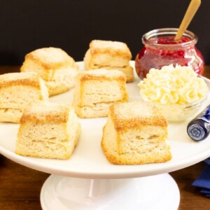 Horizontal photo of a batch of Buttermilk Sugar Biscuits on a white pedestal serving plate with butter and raspberry jam.