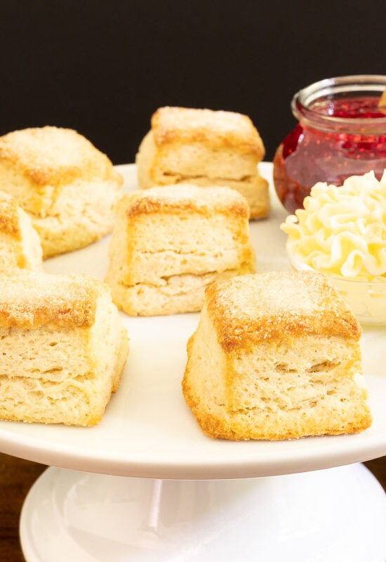 Horizontal photo of a batch of Buttermilk Sugar Biscuits on a white pedestal serving plate with butter and raspberry jam.