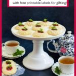 Vertical photo of a batch of Almond Shortbread Thumbprints cookies on a white pedestal serving plate surrounded by cups of tea and garnished with fresh herbs.