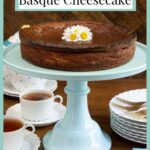 Vertical photo of a Ridiculously Easy Basque Cheesecake on a turquoise pedestal cake stand garnished with daisies.