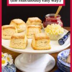 Vertical photo of a batch of Buttermilk Sugar Biscuits on a white pedestal serving plate with Raspberry Jam and butter.