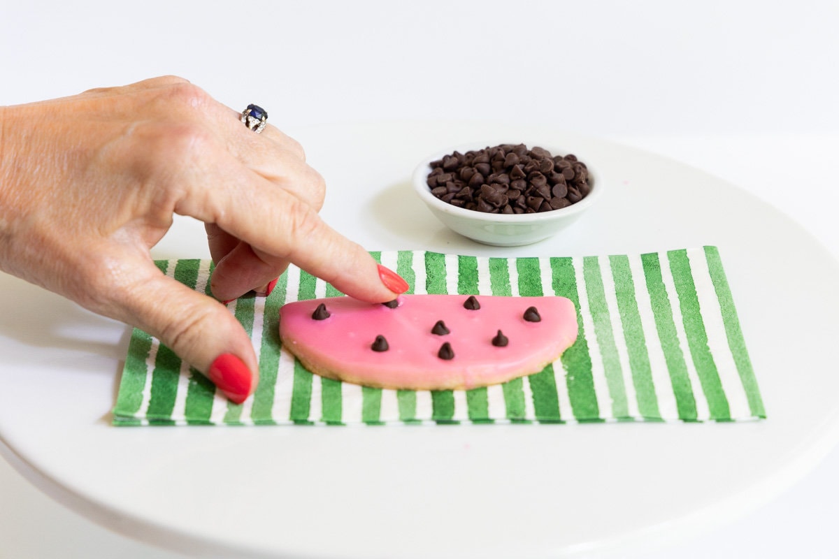Horizontal How-to photo of a person pressing the chocolate chips (watermelon "seeds") into a Watermelon Shortbread Cookie.
