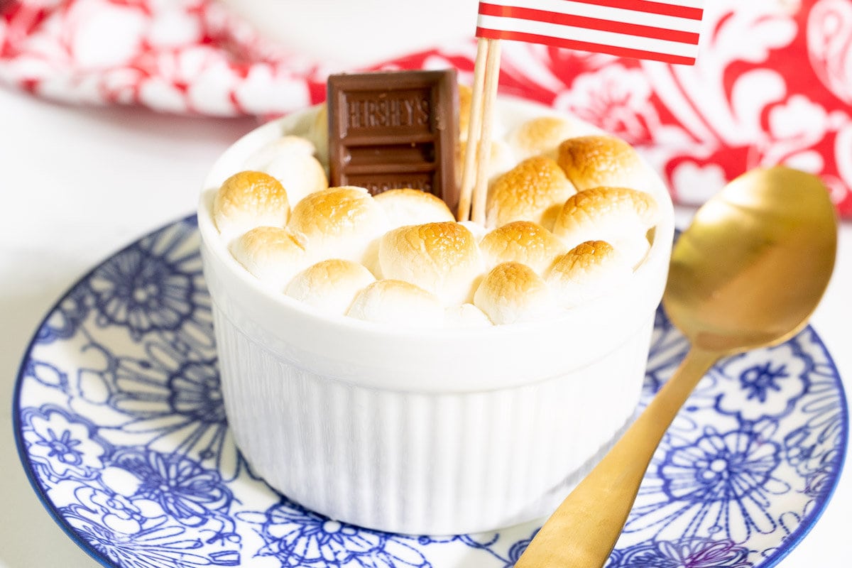 Horizontal closeup photo of a Easy Brownie S'Mores in a ramekin on a blue and white patterned plate garnished with a mini Hershey chocolate bar and an American Flag.