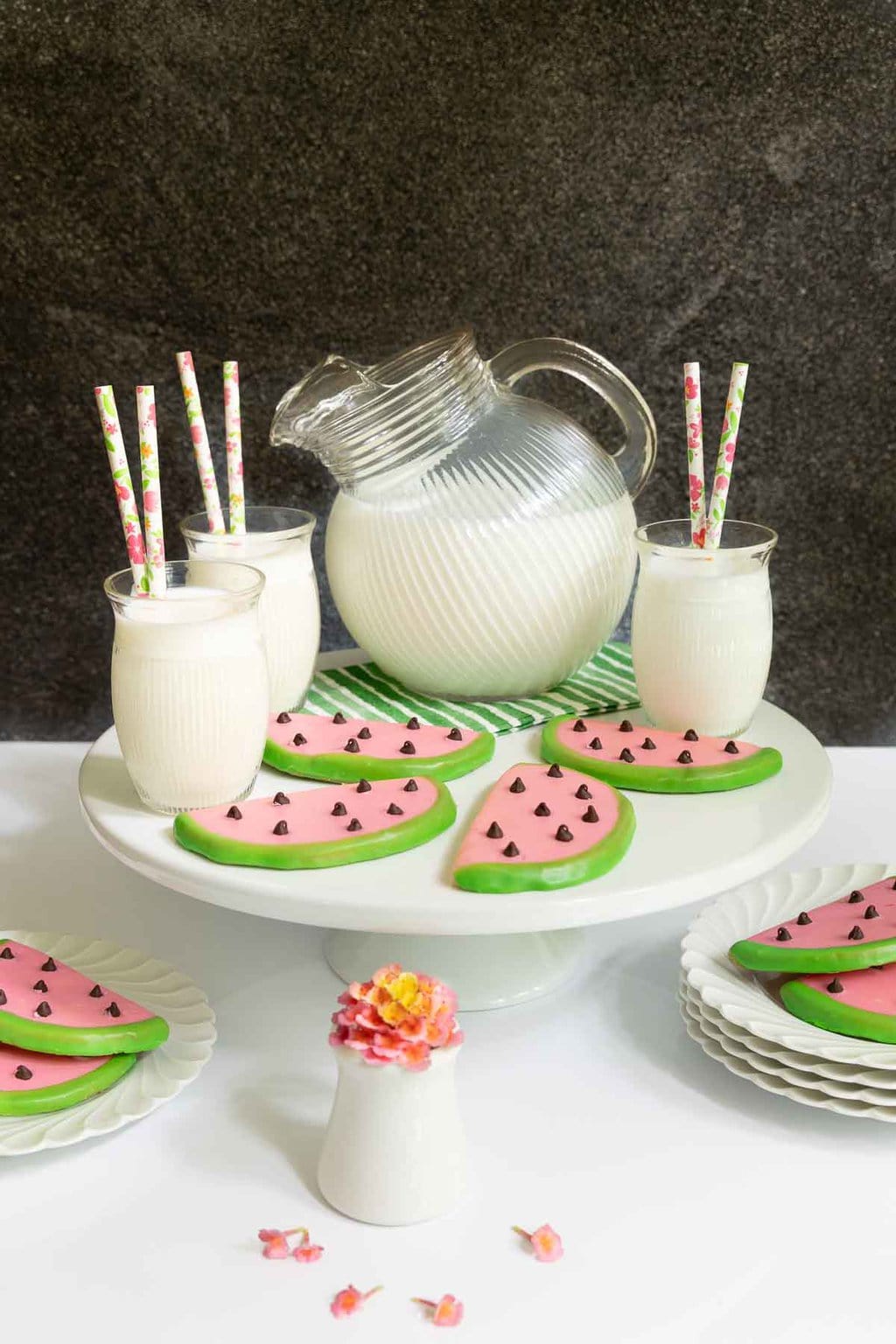 Vertical photo of a batch of Watermelon Shortbread Cookies on a white pedestal plate surrounded by glasses and a pitcher of milk.