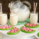 Horizontal photo of a batch of Watermelon Shortbread Cookies on a white pedestal serving plate surrounded by glasses of milk.