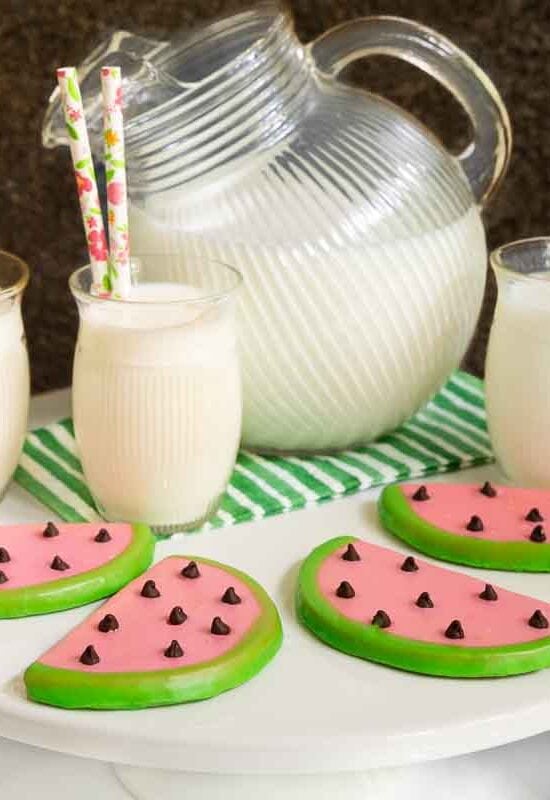 Horizontal photo of a batch of Watermelon Shortbread Cookies on a white pedestal serving plate surrounded by glasses of milk.