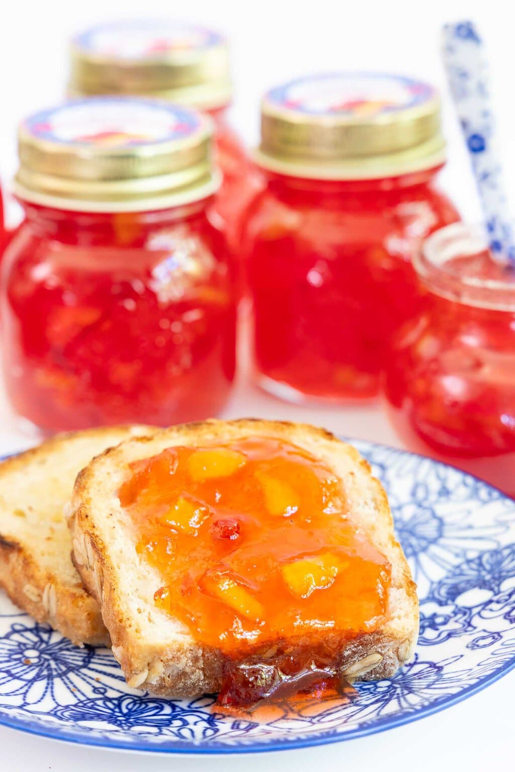 Vertical closeup photo of a slice of toast with Fresh Nectarine Jam spread over the top and jars of the jam in the background.