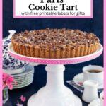 Vertical photo of a Paris Ritz Cookie Tart on a white pedestal serving plate banded with pink ribbon with polka dots and surrounded by cups of tea.