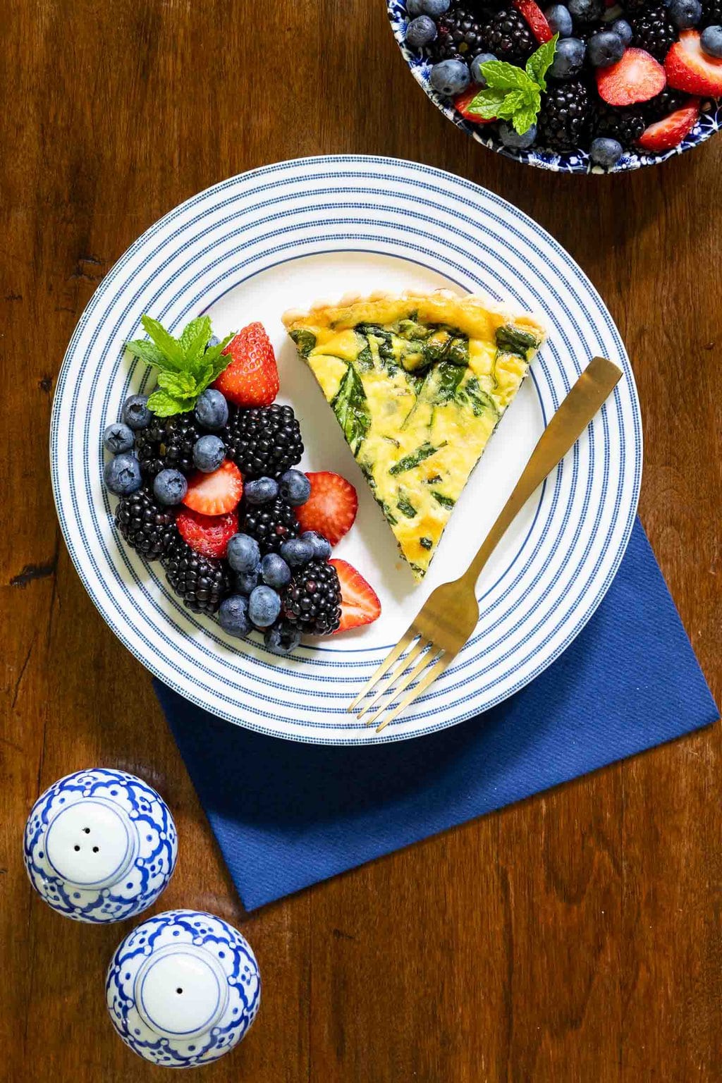 Vertical overhead photo of a French Bistro Spinach Quiche on a wood table with a side of fresh fruit.