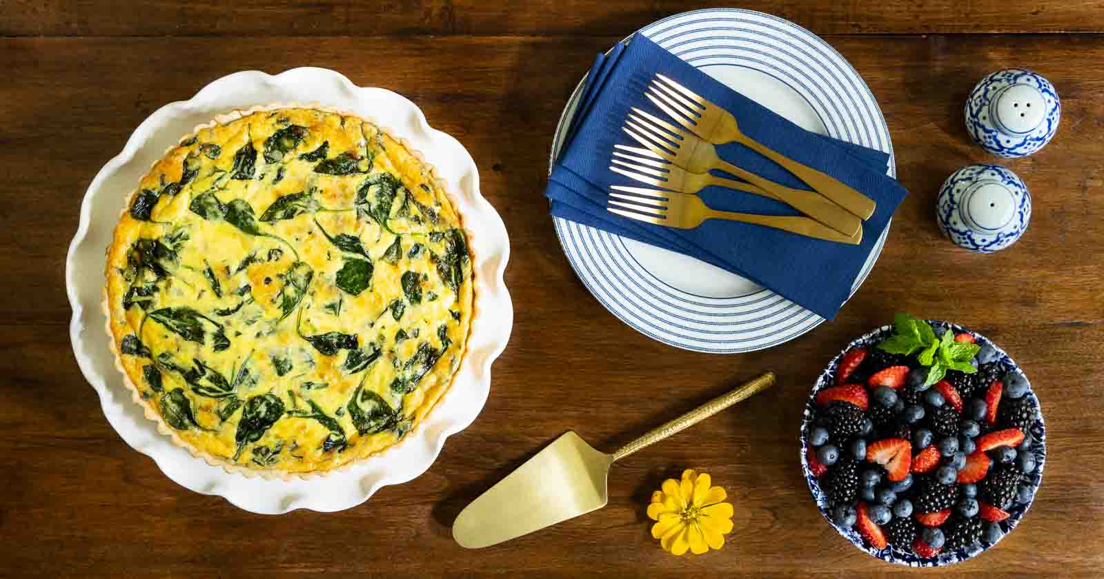 Overhead photo of a French Bistro Spinach Quiche on a scalloped white serving plate on wood table with a bowl of fresh fruit.
