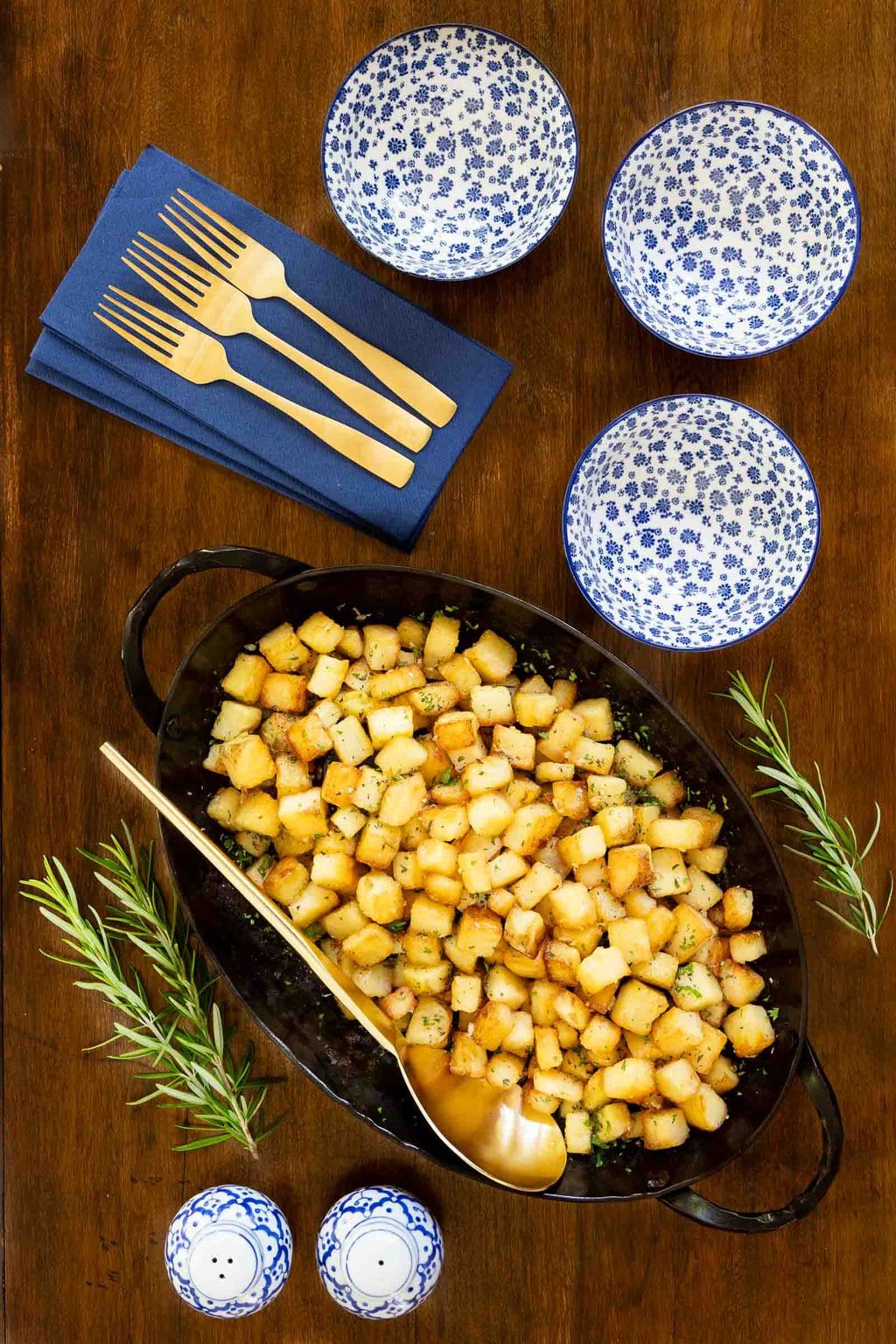 Vertical overhead photo of a batch of Rosemary Roasted French Parmentier Potatoes in an oval hammered steel serving pan on a wood table.