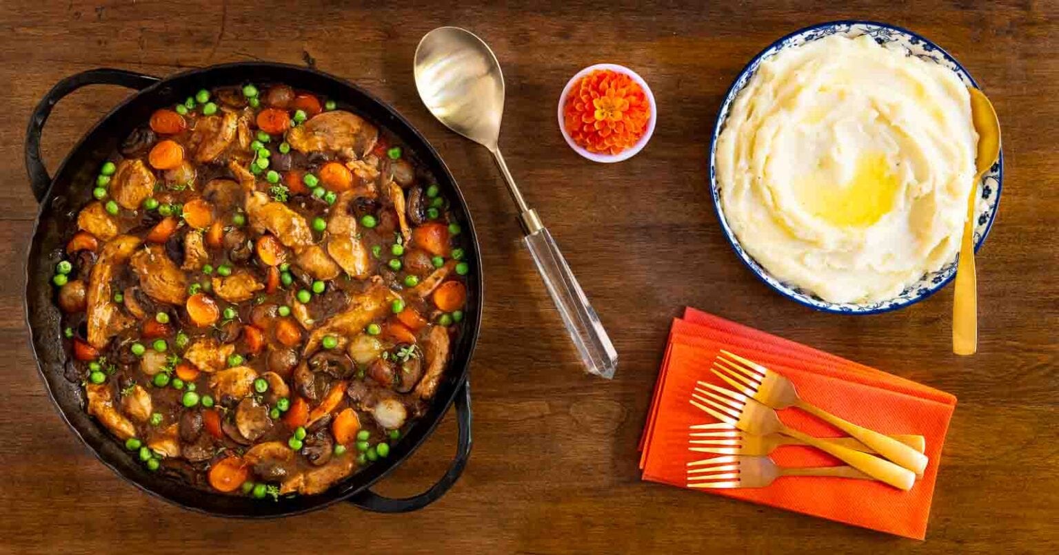Easy, Make-Ahead Parisian Coq au Vin - thecafesucrefarine.com Horizontal overhead photo of a black cast iron skillet of Easy, Make-Ahead Parisian Coq au Vin with a side of mashed potatoes on a wood table.