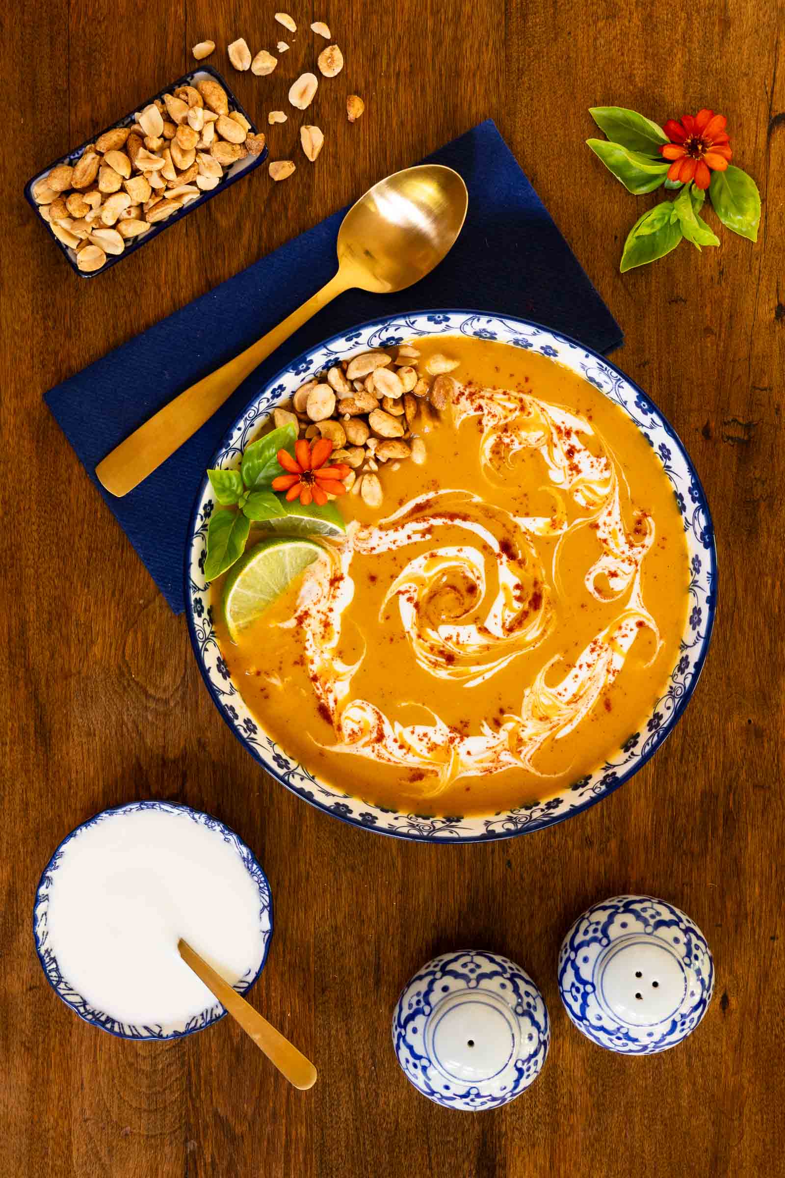 Vertical overhead photo of a bowl of Roasted Sweet Potato Soup garnished with sour cream, basil leaves, peanuts and lime wedges.