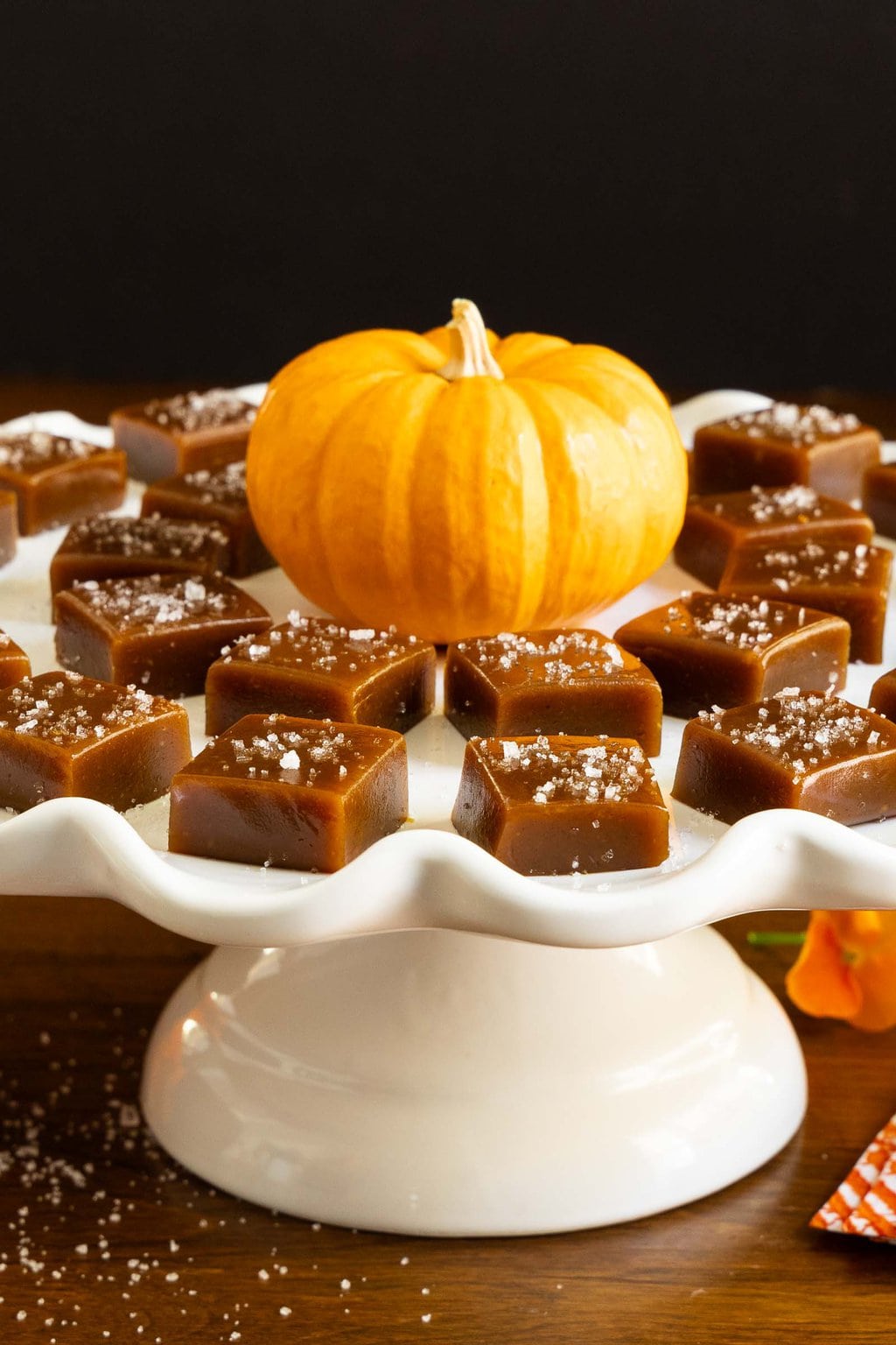 Vertical photo of a white pedestal serving plate filled with Ridiculously Easy Sea-Salted Pumpkin Microwave Caramels.
