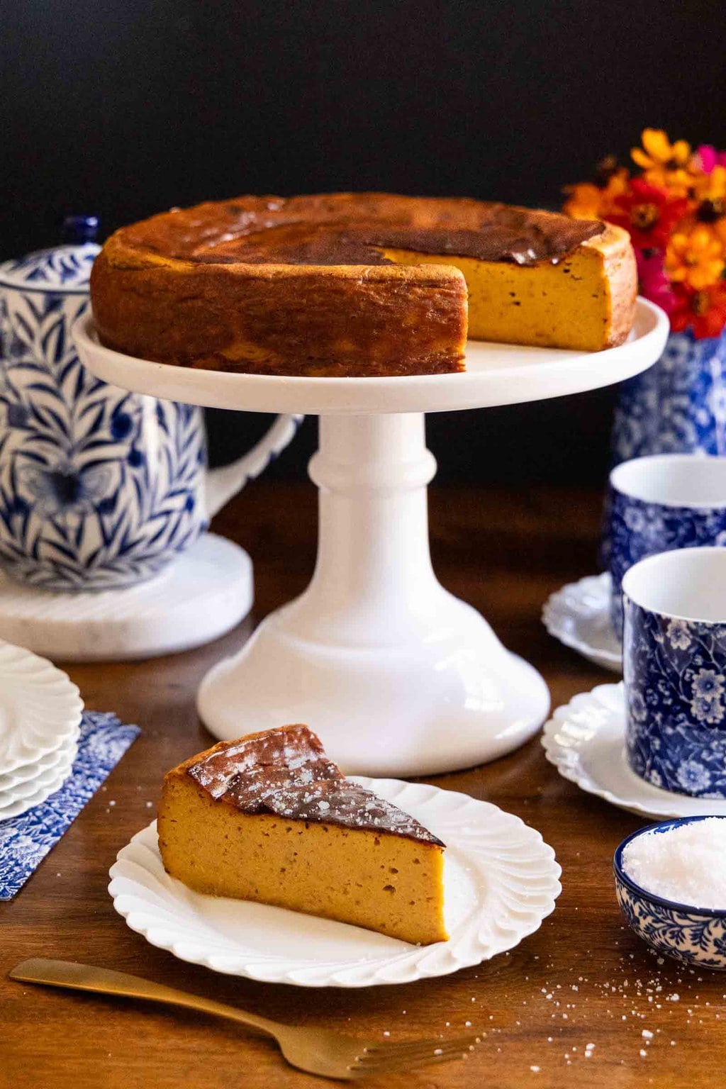 Vertical photo of a Ridiculously Easy Basque Sweet Potato Cheesecake on a white pedestal serving plate with an individual slice on a white serving plate in the foreground.