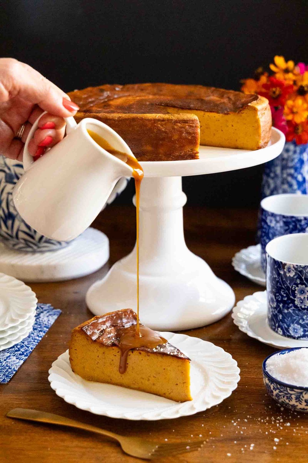 Vertical photo of a person pouring warm caramel sauce over a slice of Ridiculously Easy Basque Sweet Potato Cheesecake with the whole cheesecake in the background.
