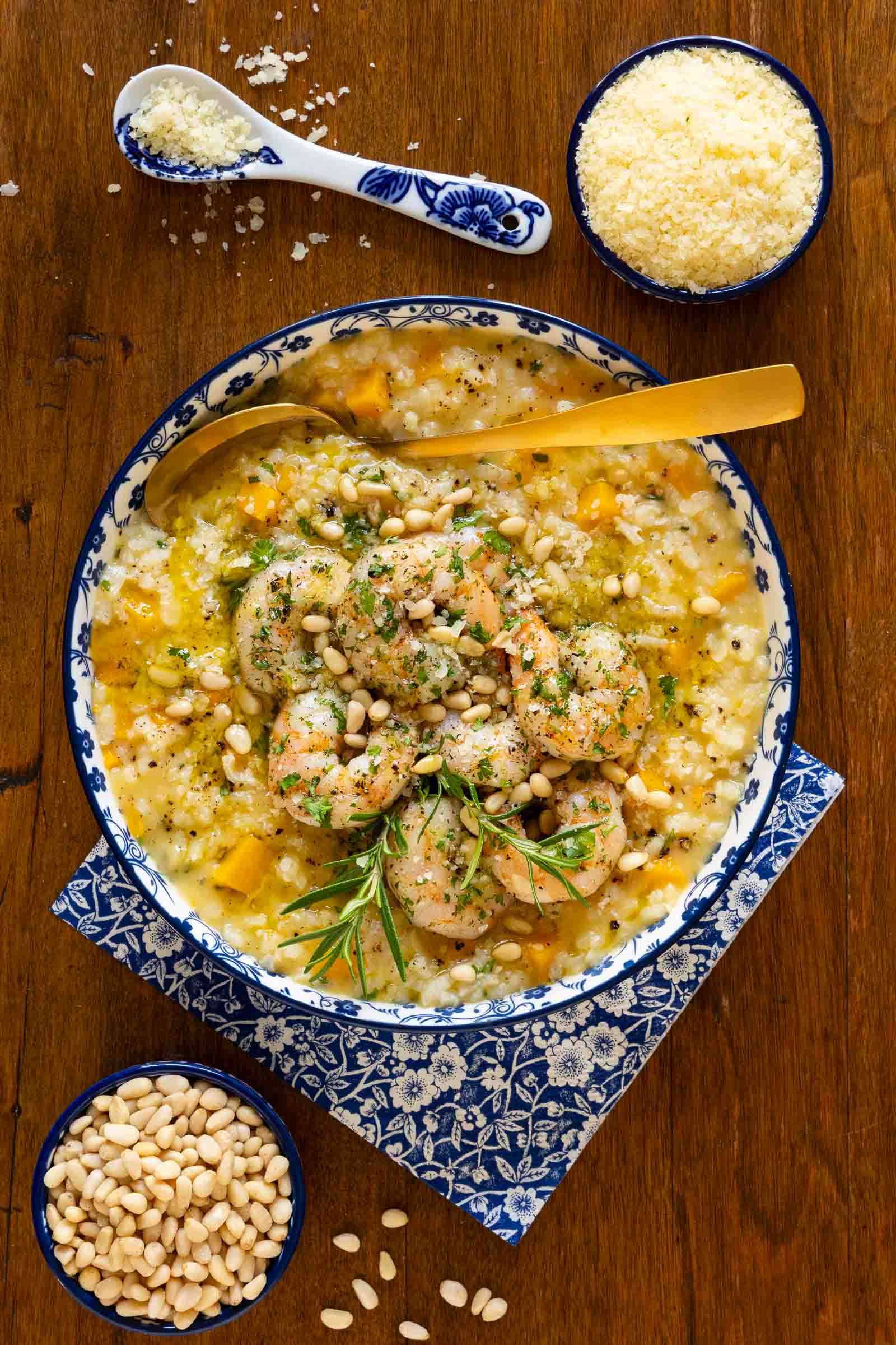 Vertical overhead photo of a serving bowl of Butternut Squash Risotto with Italian Roasted Shrimp on a wood table with bowls of grated Parmesan cheese and pine nuts.
