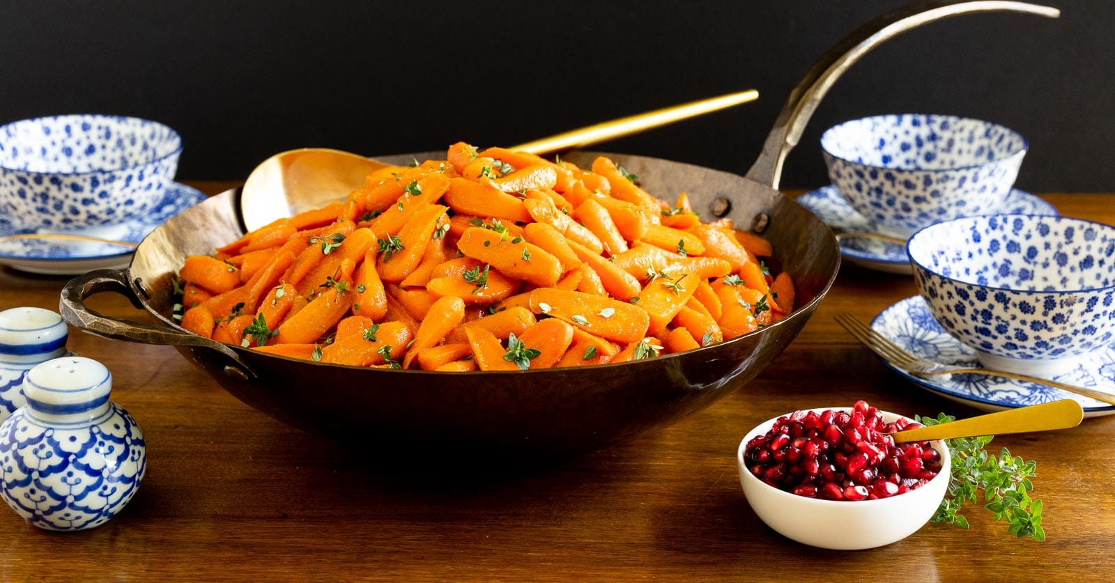 Horizontal photo of a hammered steel pan of Stove-top Honey Glazed Carrots on a wood table with a side of pomegranate arils.
