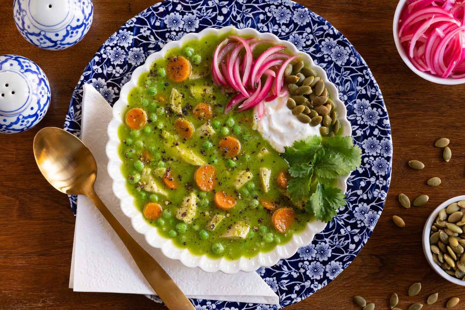 Overhead horizontal photo of a bowl of Peruvian Chicken and Rice Soup in a white scalloped serving bowl and garnished with pickled red onions, pepitas, yogurt and cilantro leaves.
