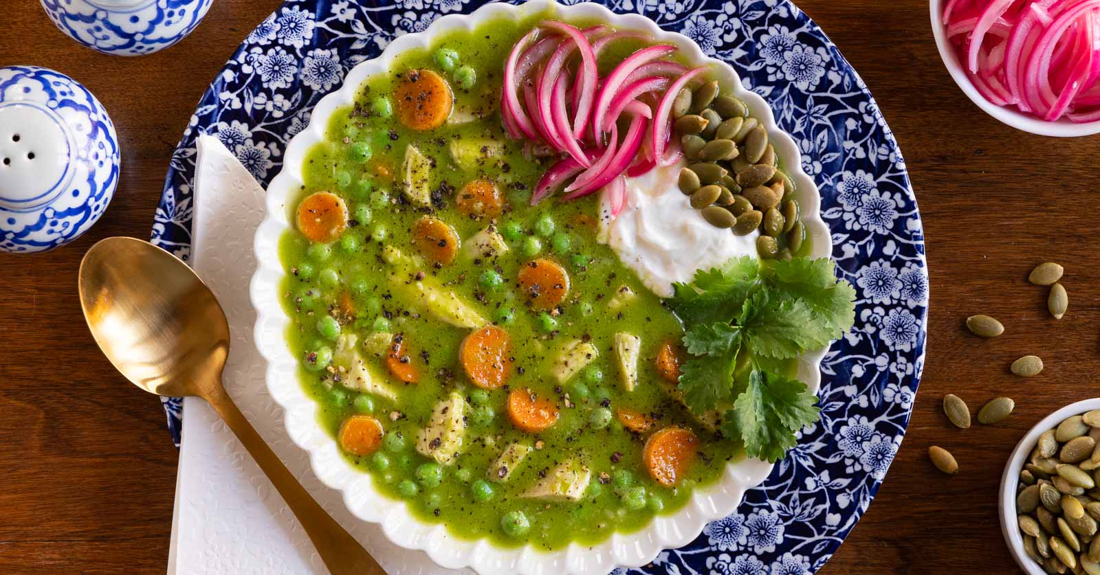 Horizontal overhead photo of a bowl of Peruvian Chicken and Rice Soup in a white serving bowl on a wood table garnished with cilantro, pepitas, pickled red onions and yogurt.