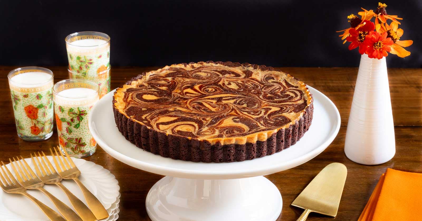 Horizontal photo of a Ridiculously Easy Caramel Cream Cheese Tart on a white pedestal serving plate surrounded by dinnerware and glasses of milk.