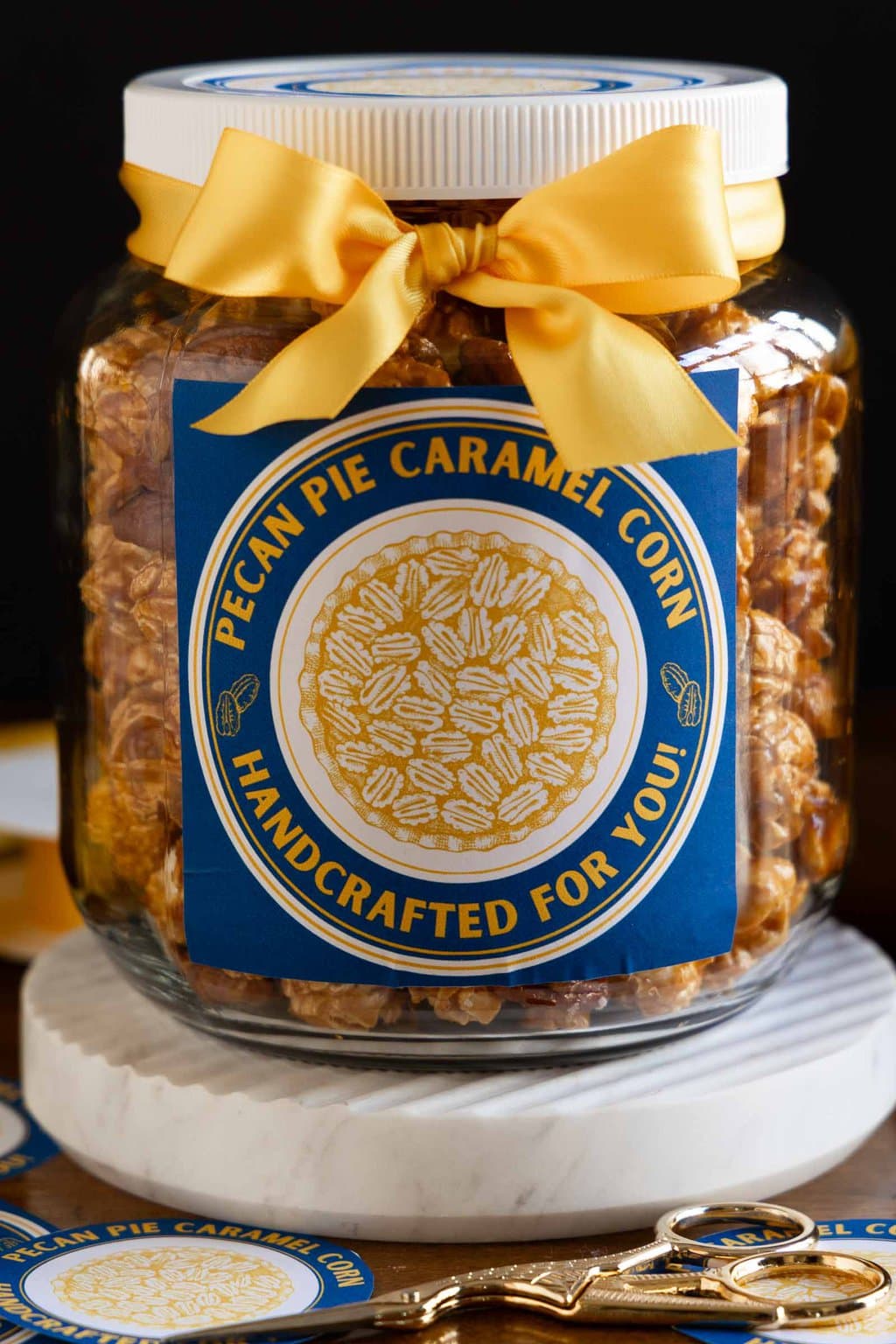 Vertical closeup photo of a plastic lidded jar filled with Ridiculously Easy Pecan Pie Caramel Corn and decorated with a gold bow and a custom label for gift giving.