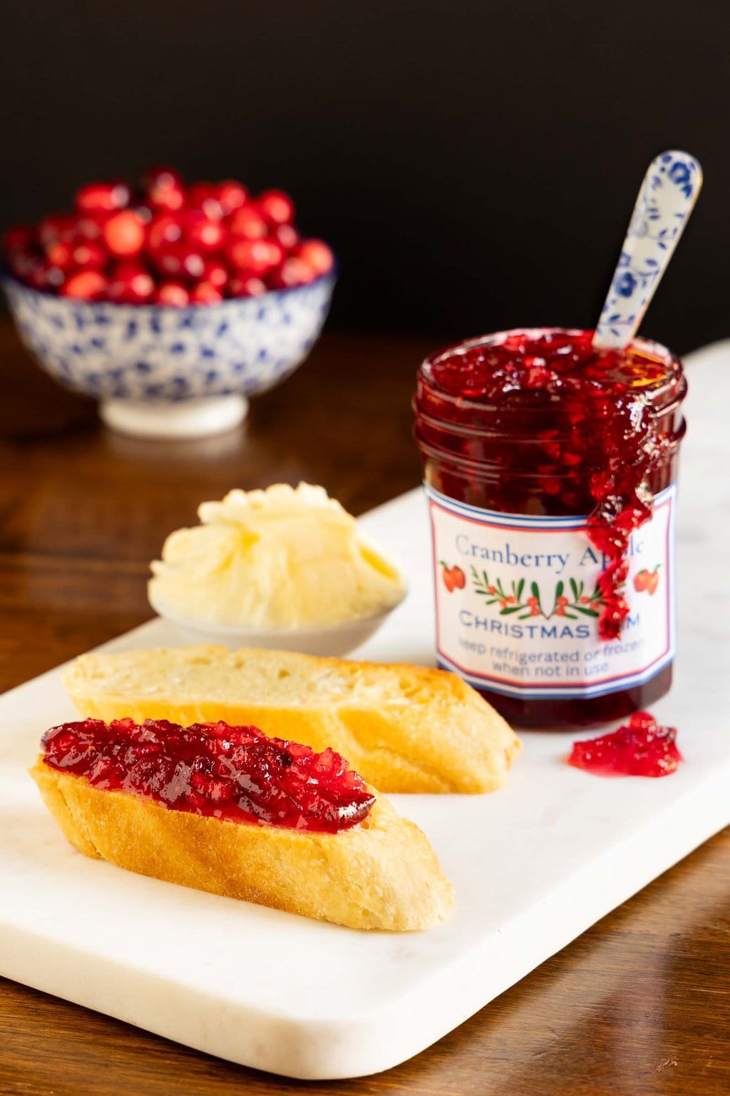 Vertical photo of a jar of Cranberry Apple Christmas Jam next to slices of bread with the jam on top.