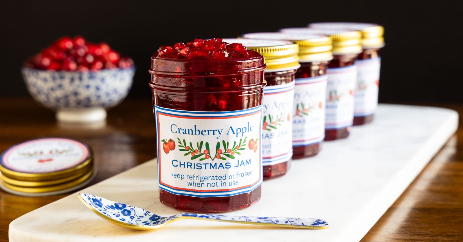 Horizontal closeup photo of a row of jars of Cranberry Apple Christmas Jam on a white marble slab.