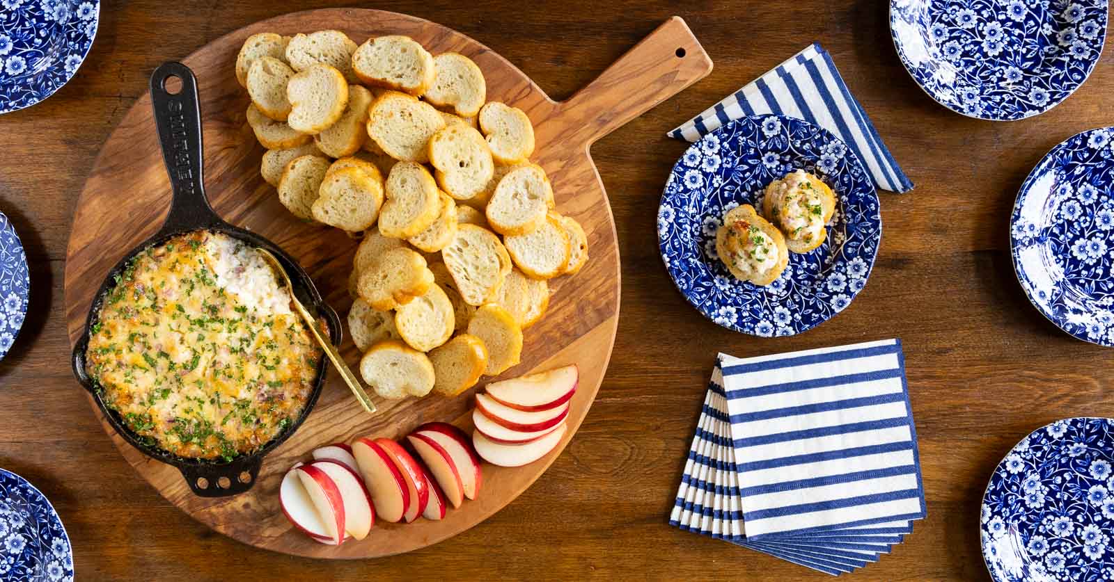 Overhead horizontal photo of a cast iron skillet of Ridiculously Easy Hot Onion Dip surrounded by sliced apples and crostini.