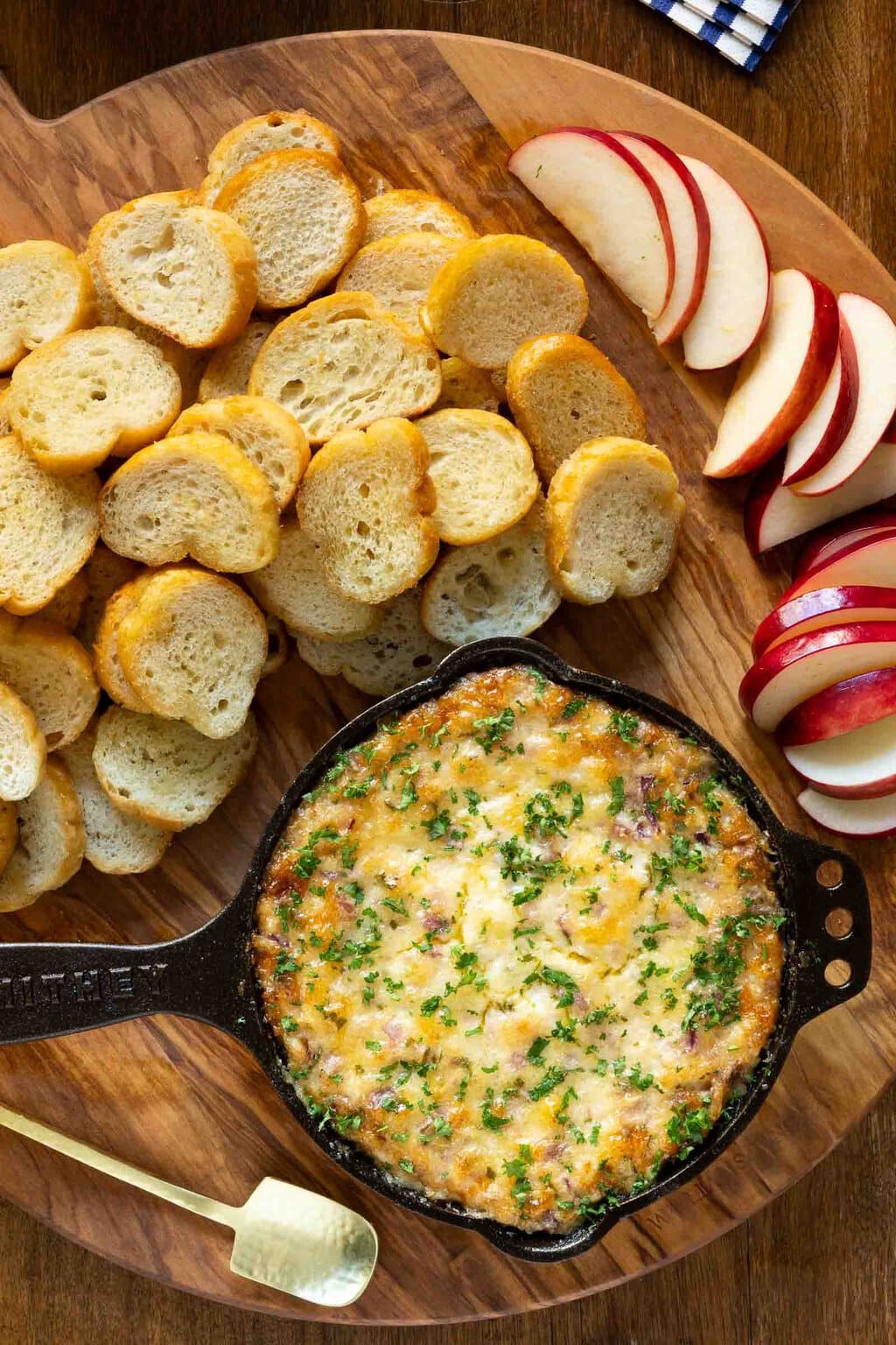 Vertical overhead photo of a black cast iron pan of Ridiculously Easy Hot Onion Dip on a wood appetizer tray surrounded by slices of apples and crostini.