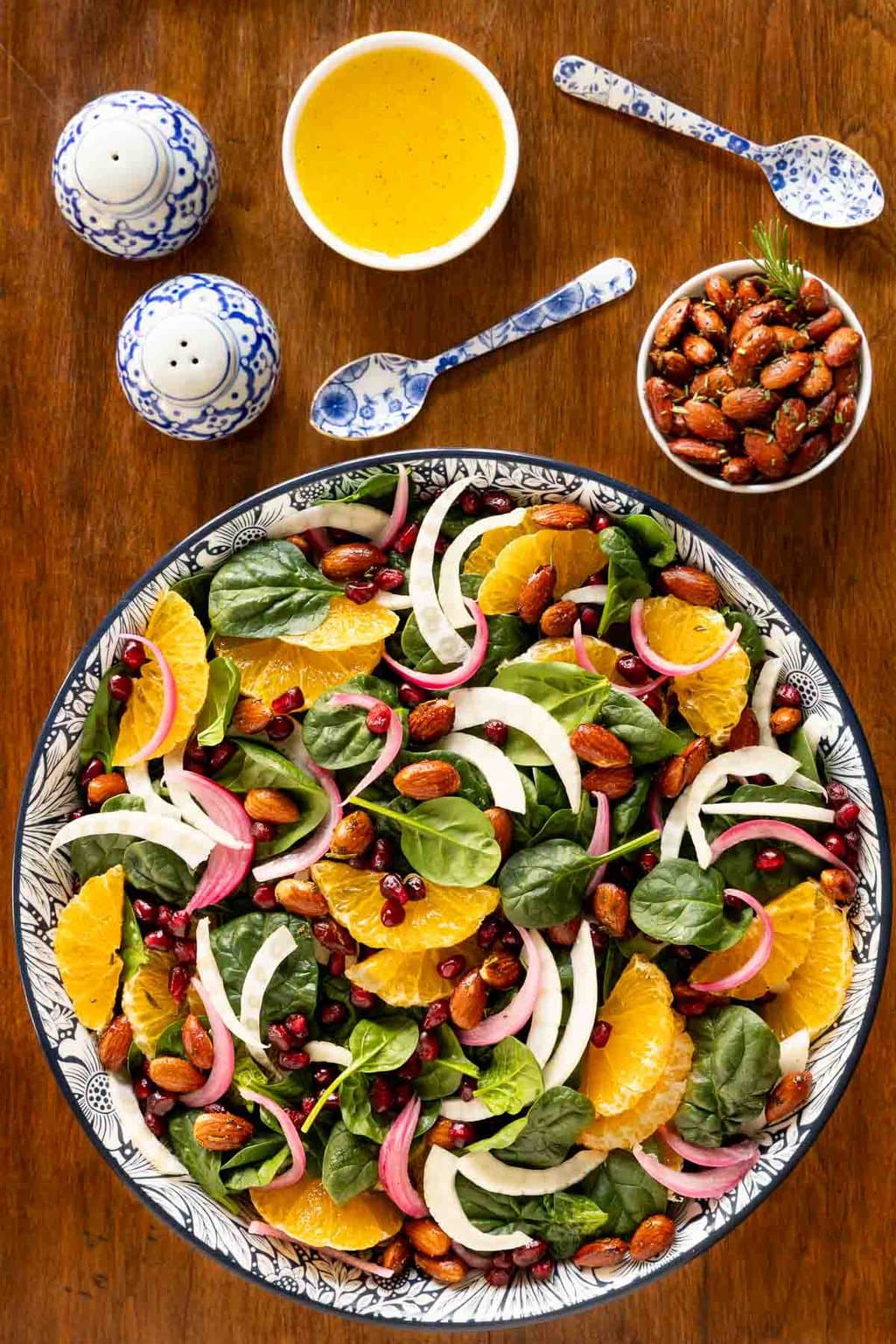 Vertical overhead photo of a bowl of Orange Fennel Spinach Salad with Honey White Balsamic Dressing on a wood table with bowls of dressing and almonds.