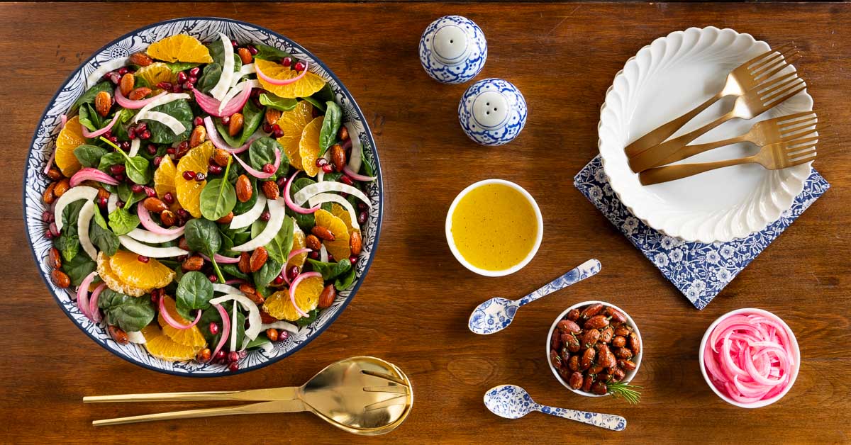 Overhead horizontal photo of a bowl of Orange Fennel Spinach Salad with Honey White Balsamic Dressing surrounded by small bowls of the dressing, roasted almonds and pickled onions.