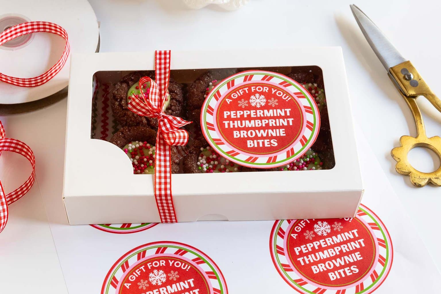 Horizontal photo of a windowed gift box filled with Peppermint Thumbprint Brownie Bites with a custom label for gift giving on the top of the box.