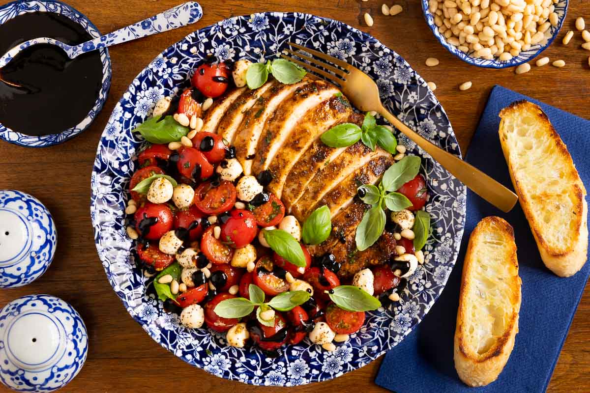 Sliced pan-seared chicken breast served with cherry tomatoes, mozzarella pearls, fresh basil, pine nuts, and balsamic glaze on a blue floral plate with toasted bread alongside.