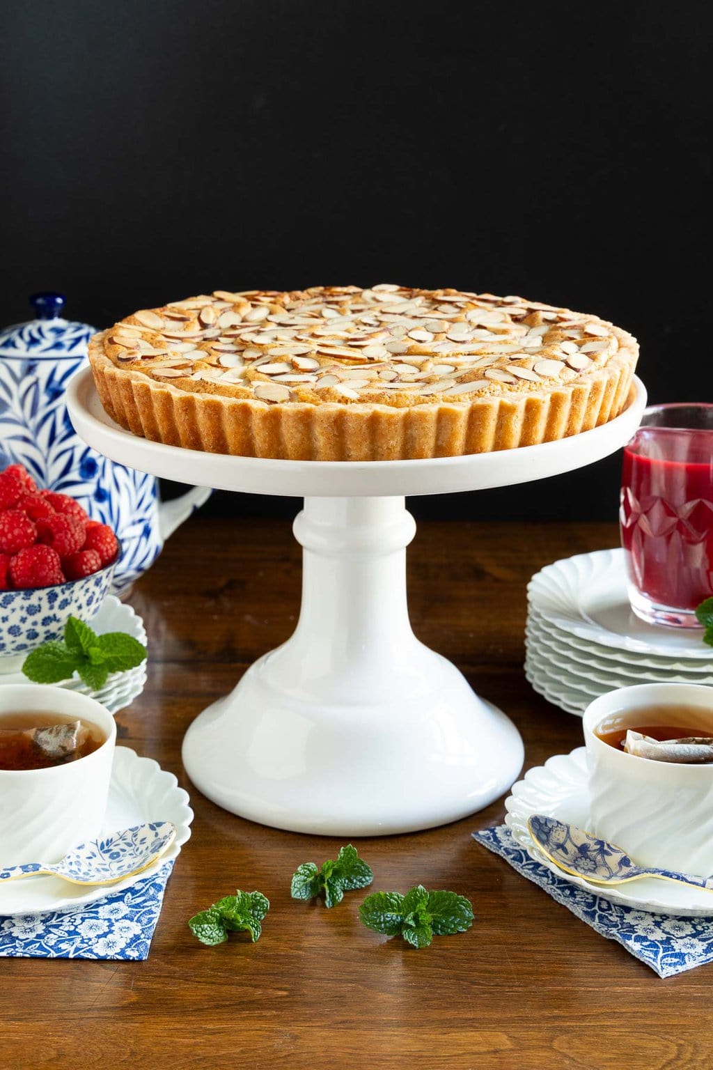 Vertical photo of a Ridiculously Easy French Almond Frangipane Tart on a white pedestal serving plate surrounded by raspberry coulis and cups of tea.