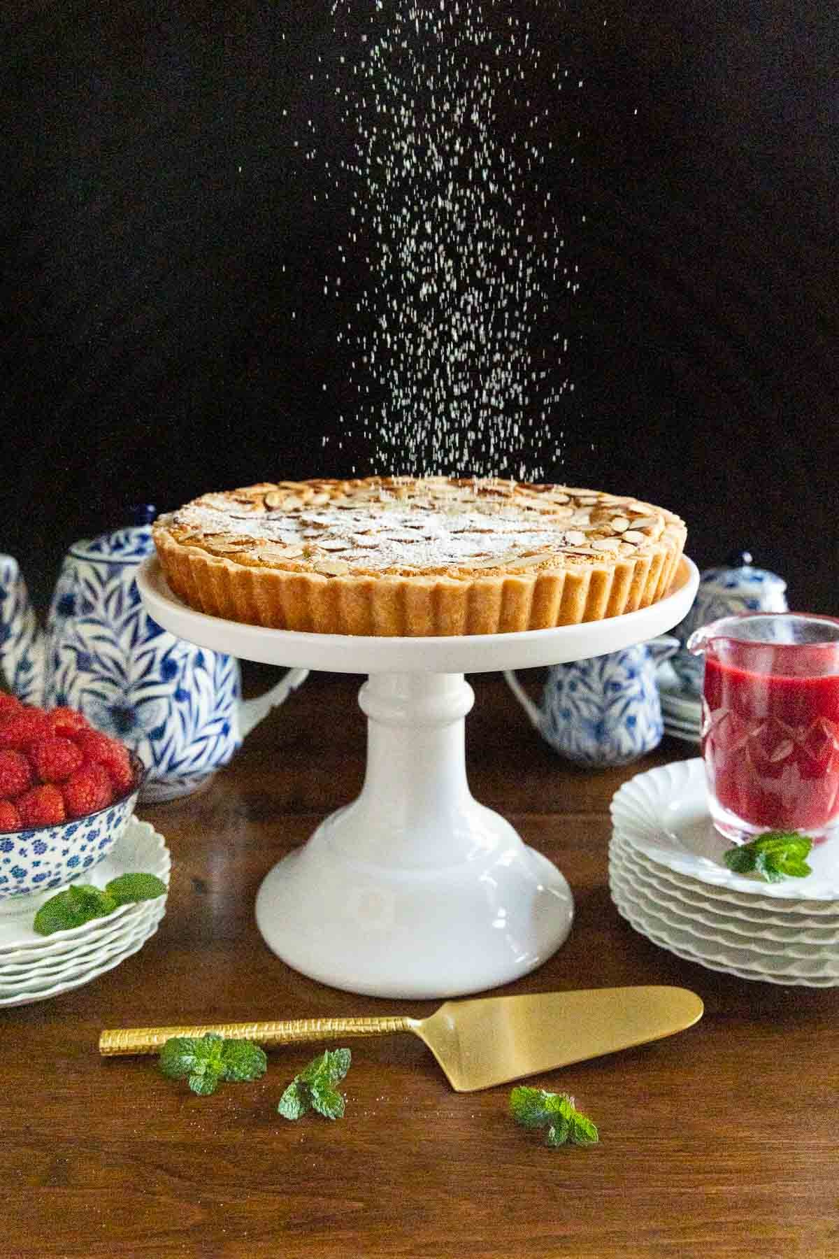 Vertical photo of a Ridiculously Easy French Almond Frangipane Tart on a white pedestal serving plate being sprinkled with powdered sugar.