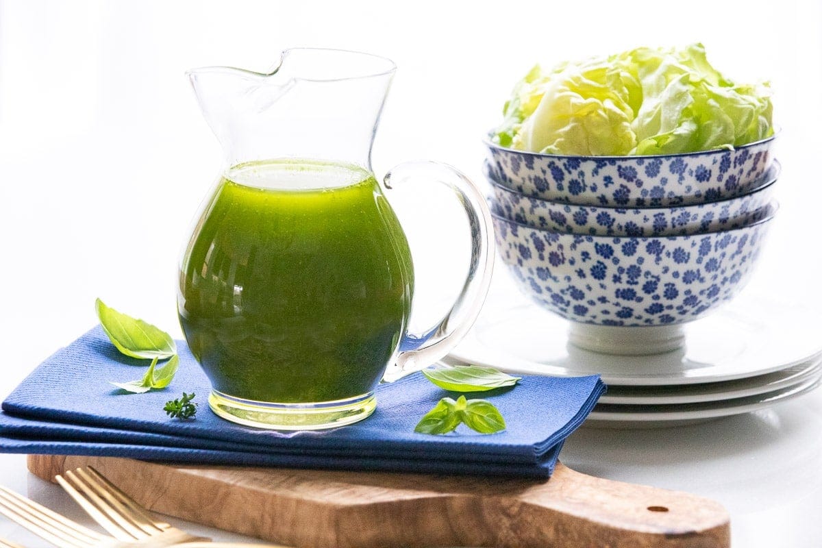 Glass pitcher of bright green Greek herb dressing with fresh basil on a blue napkin, stacked blue patterned bowls filled with butter lettuce in the background.