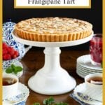 Vertical photo of a French Almond Frangipane Tart on a white pedestal serving plate surrounded by fresh raspberries, mint leaves and a glass pitcher of Raspberry Coulis.