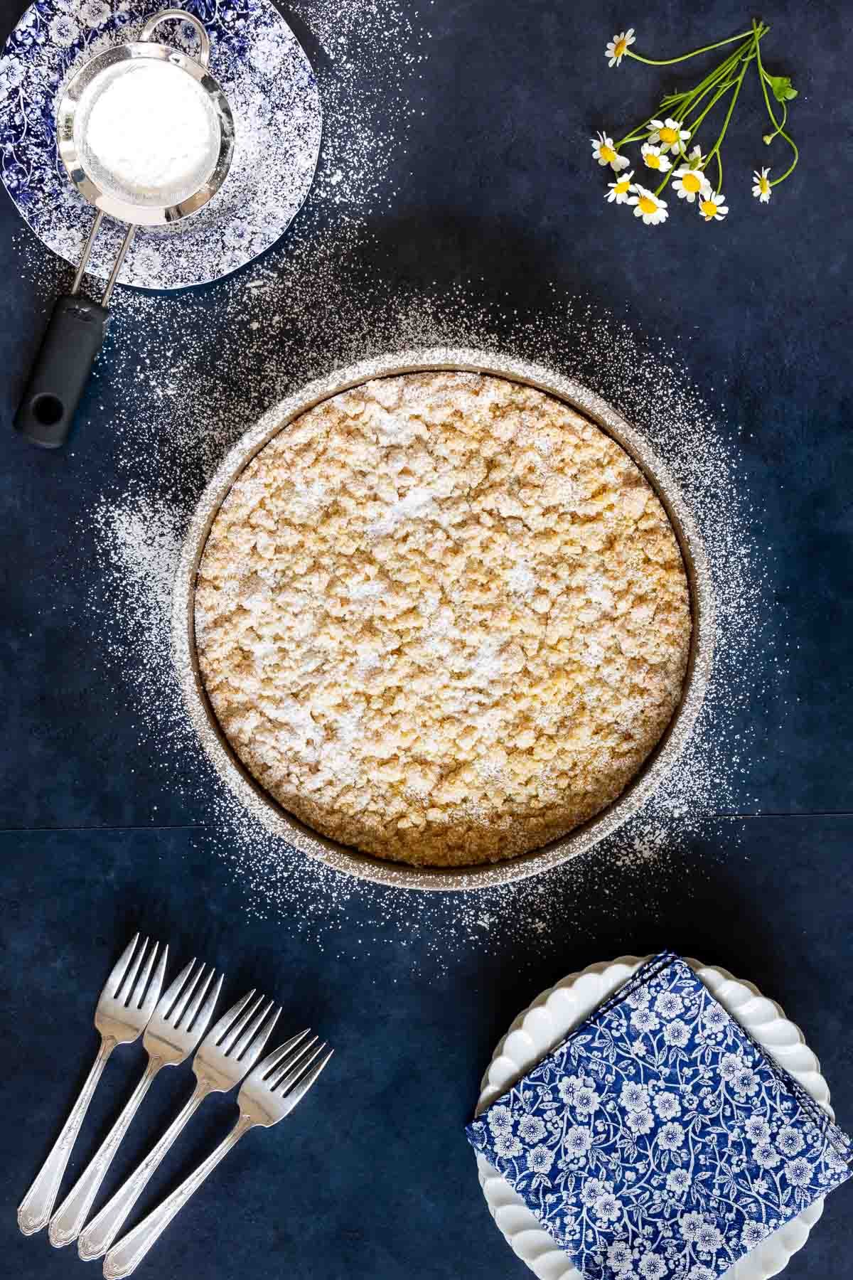 Vertical overhead photo of a Italian Lemon Curd Ricotta Crumb Cake on a dark blue surface and sprinkled with powdered sugar.